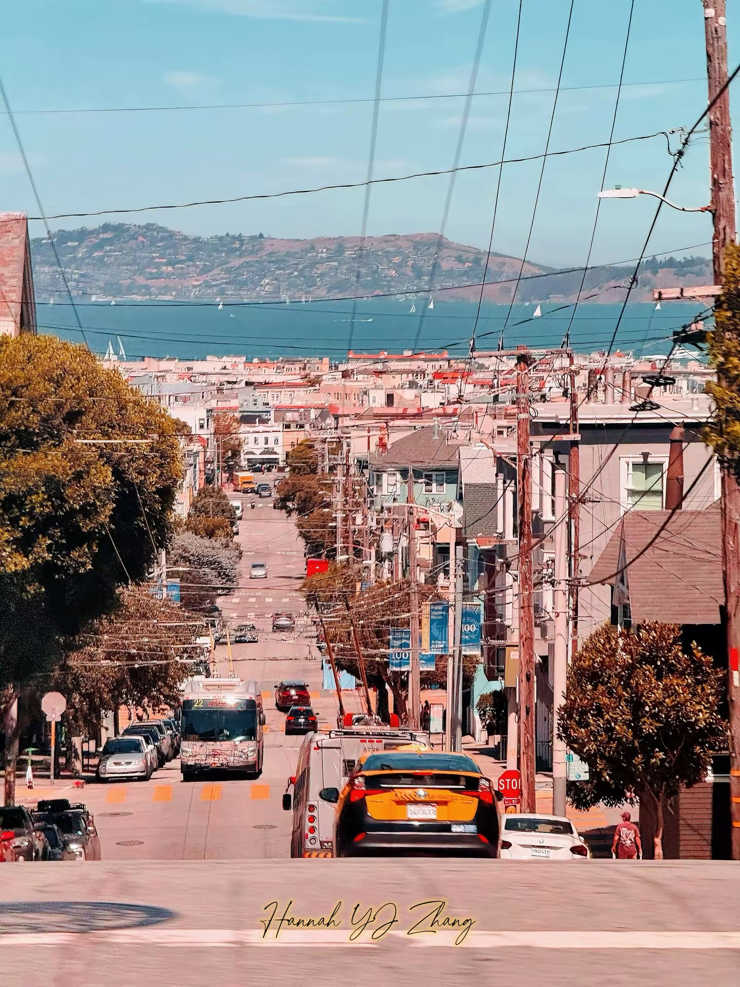 A steep city street descending toward a bay filled with sailboats and a hillside beyond